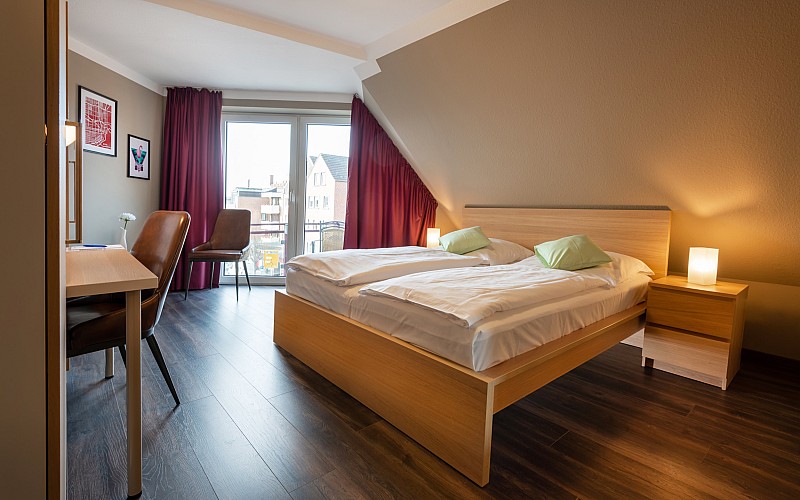 Modern eingerichtetes Hotelzimmer mit zwei Einzelbetten, Schreibtisch, Fernseher, Sitzbereich und bodentiefem Fenster.